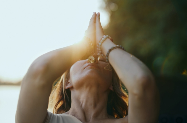 Person hält Hände in Namaste-Pose zum Himmel. Im Hintergrund geht die Sonne unter.