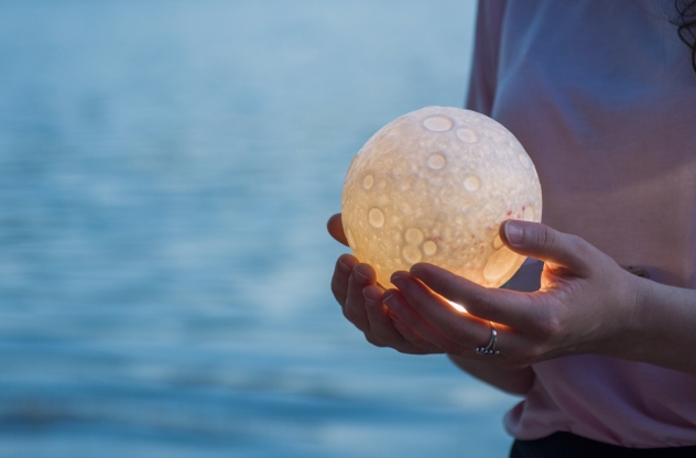 Person hält einen kleinen Leucht-Mond in den Händen. Im Hintergrund ist Wasser zu sehen.