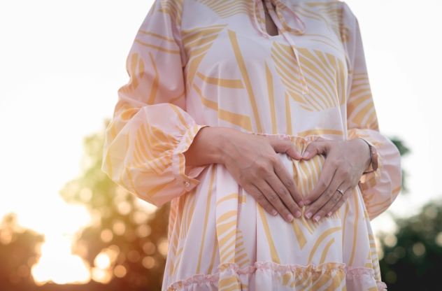 Frau in luftigem Sommerkleid umarmt ihren Bauch und formt ein Herz und hat einen unerfüllten Kinderwunsch