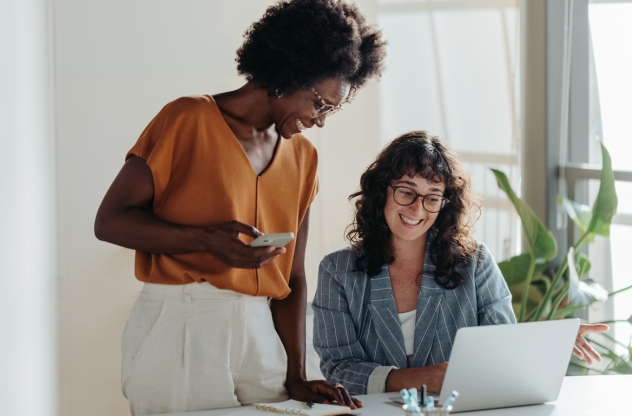Zwei berufstätige Frauen lächeln und arbeiten gemeinsam an einem Laptop in einem hellen Büro.
