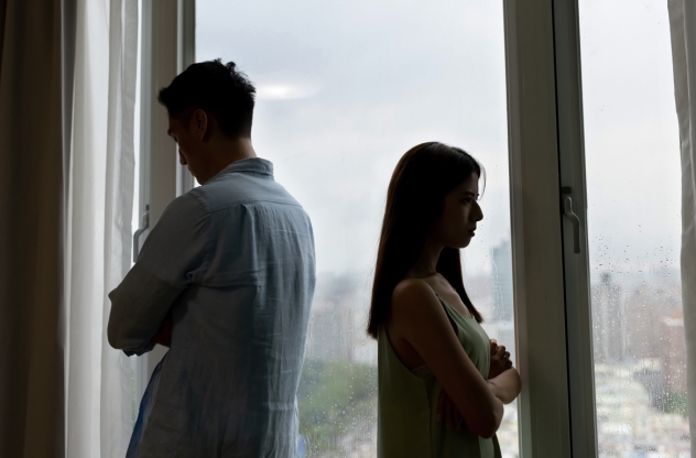 Ein Mann und eine Frau stehen abgewandt voneinander vor einem Fenster, eine sichtbare Spannung zwischen ihnen. Trennung nach langer Beziehung