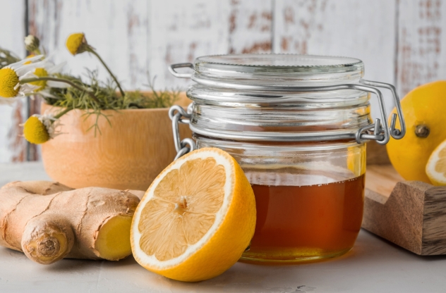 Gesund durch die Erkältungszeit: Ein Glas Honig neben einer halben Zitrone und einem Stück Ingwer, arrangiert auf einer rustikalen Holzunterlage.