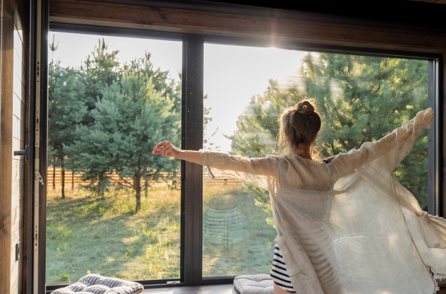 Eine Person mit ausgebreiteten Armen steht vor einem großen Fenster und genießt den Blick auf einen sonnigen Garten mit Bäumen.