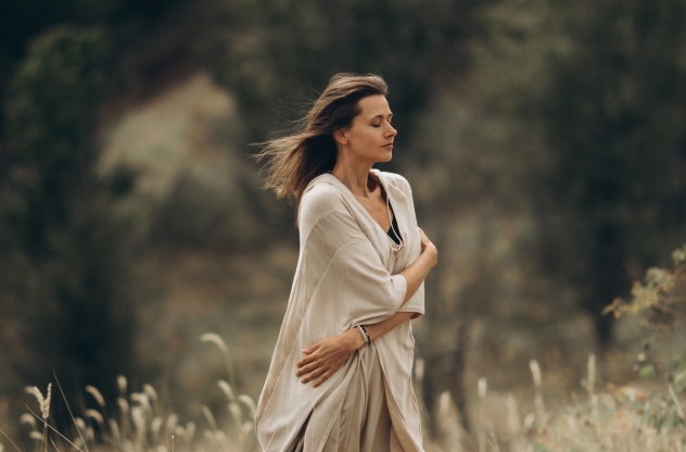 Eine Frau mit geschlossenen Augen steht in der Natur und hält ihre Arme um sich selbst geschlungen, während der Wind ihr Haar weht. | Spiritual Bypassing