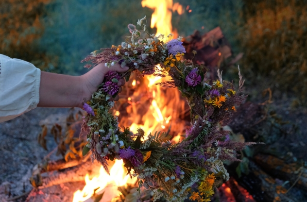 Beltane: Eine Hand hält einen bunten Blumenkranz über ein loderndes Feuer bei einem Ritual im Freien.