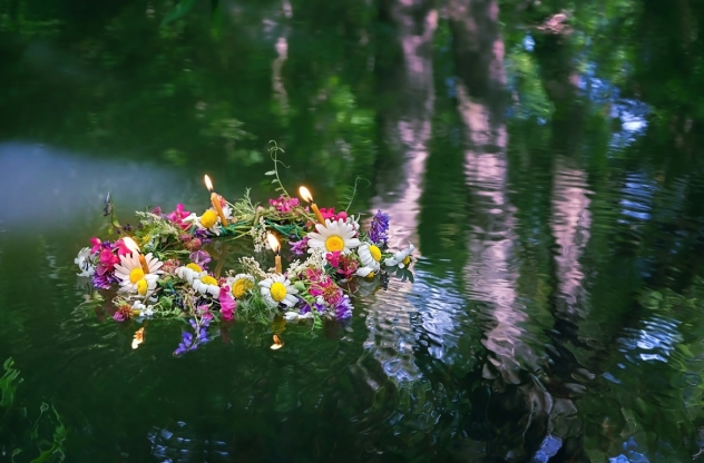 Jahreskreisfeste: Ein bunter Blumenkranz mit brennenden Kerzen schwimmt auf ruhigem Wasser im Wald.