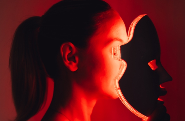 Rotlichttherapie: Eine Frau trägt eine rot leuchtende LED-Lichttherapiemaske in einem dunklen Raum.