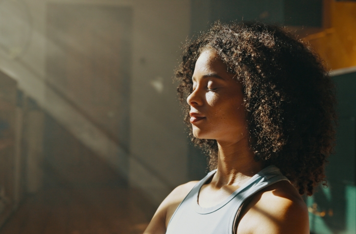 Eine junge Frau hält mit geschlossenen Augen in einem lichtdurchfluteten Raum inne und genießt einen Moment der Ruhe und des tiefen Durchatmens.