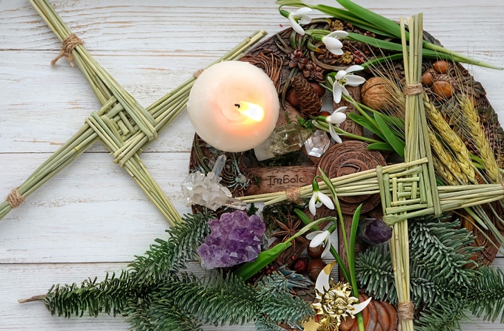 Ein Arrangement zur Feier von Imbolc mit einer brennenden Kerze, umgeben von zwei geflochtenen Brigidskreuzen aus Stroh, Tannenzweigen, Schneeglöckchen und verschiedenen Naturmaterialien.