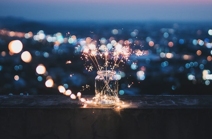 Ein Einmachglas mit brennenden Wunderkerzen steht auf einer Brüstung vor dem unscharfen, nachtblauen Lichtermeer einer Stadt. Die Szene vermittelt eine gemütliche Silvesterstimmung.