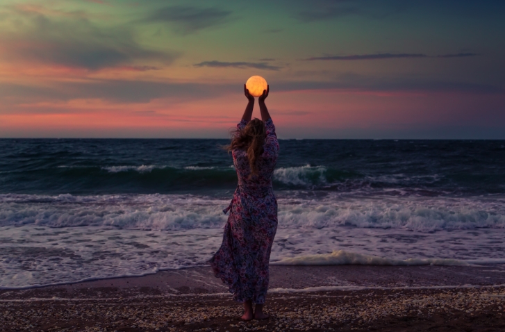 Rückansicht einer Frau im Kleid am Strand, die vor dem Vollmond ihre Arme emporstreckt | Vollmondrituale.
