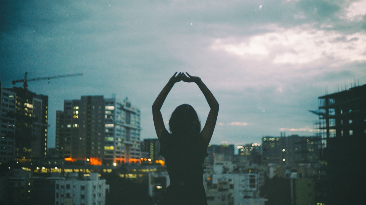 Eine Frau steht mit erhobenen Armen vor einer nächtlichen Stadtkulisse und blickt in den bewölkten Himmel.