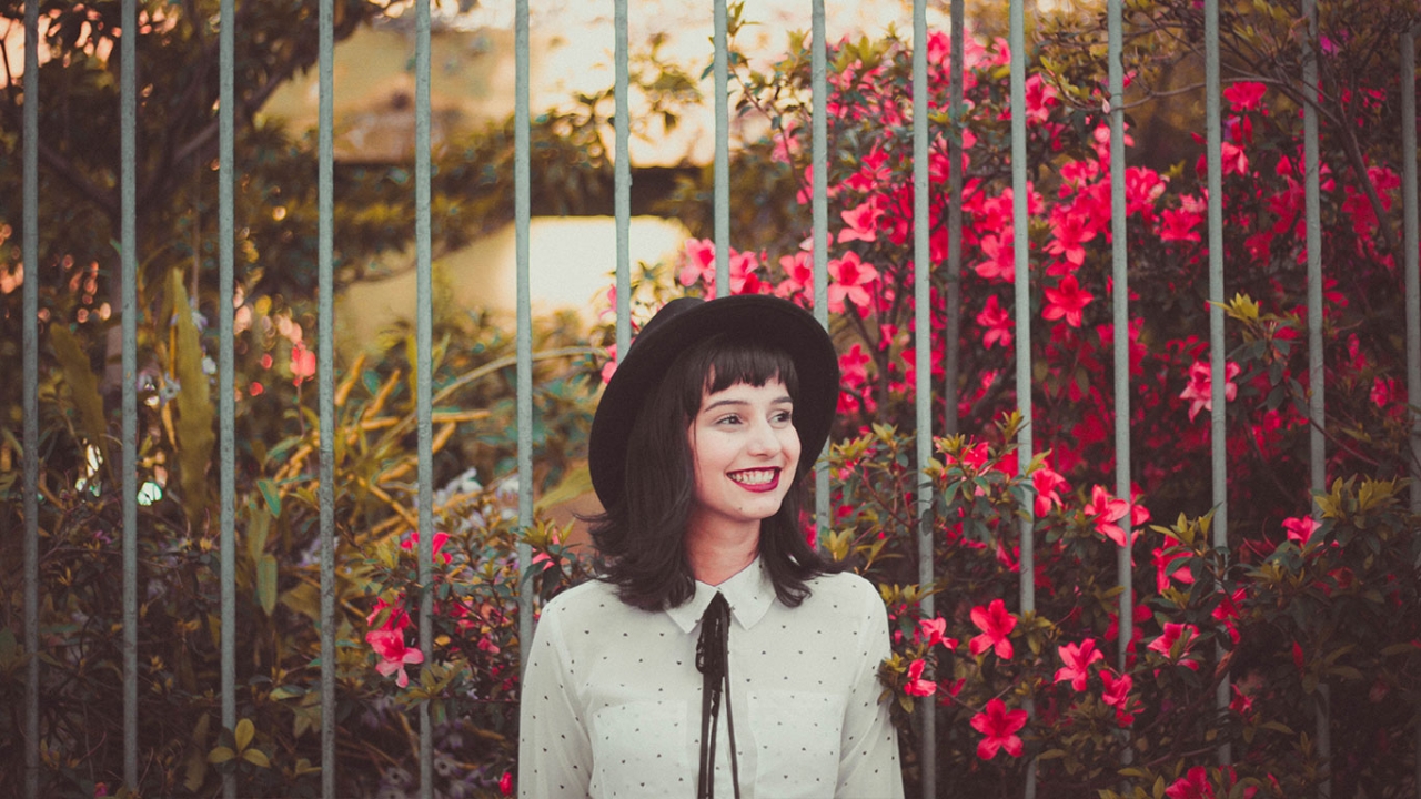 Selbstwertgefühl stärken: Eine lächelnde Frau steht vor einem Zaun und blühenden pinken Blumen