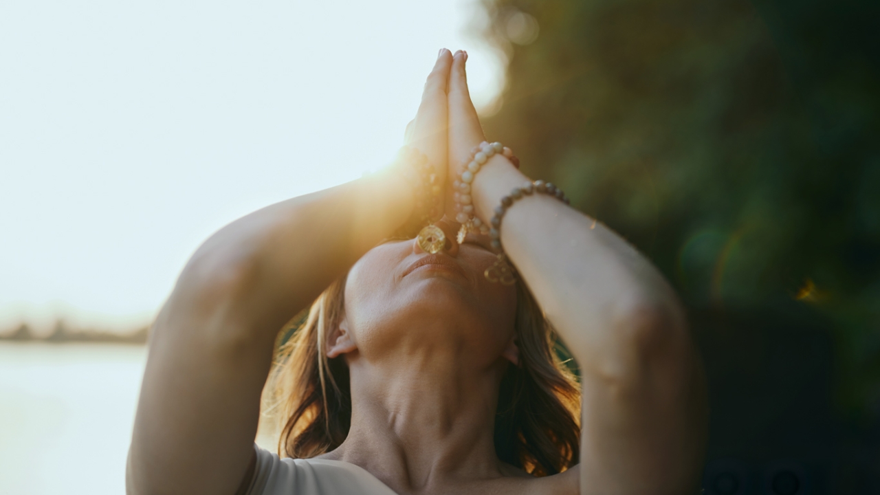 Person hält Hände in Namaste-Pose zum Himmel. Im Hintergrund geht die Sonne unter.