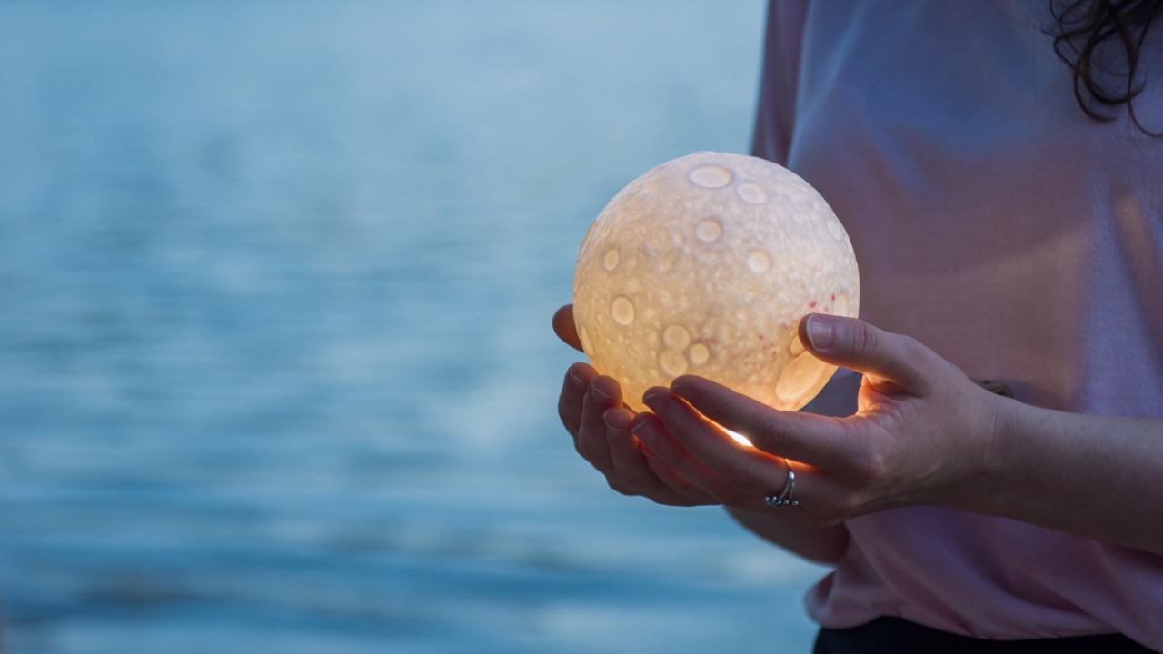 Person hält einen kleinen Leucht-Mond in den Händen. Im Hintergrund ist Wasser zu sehen.