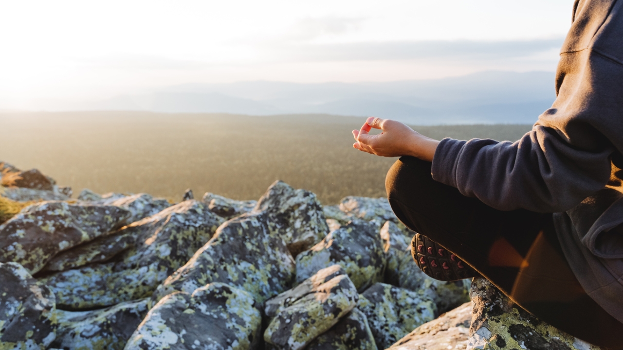 Mit 4 Hatha-Yoga-Übungen dein Wohlbefinden und deine Laune steigern Eine Frau meditiert. Im Hintergrund sieht man Felder und Berge.