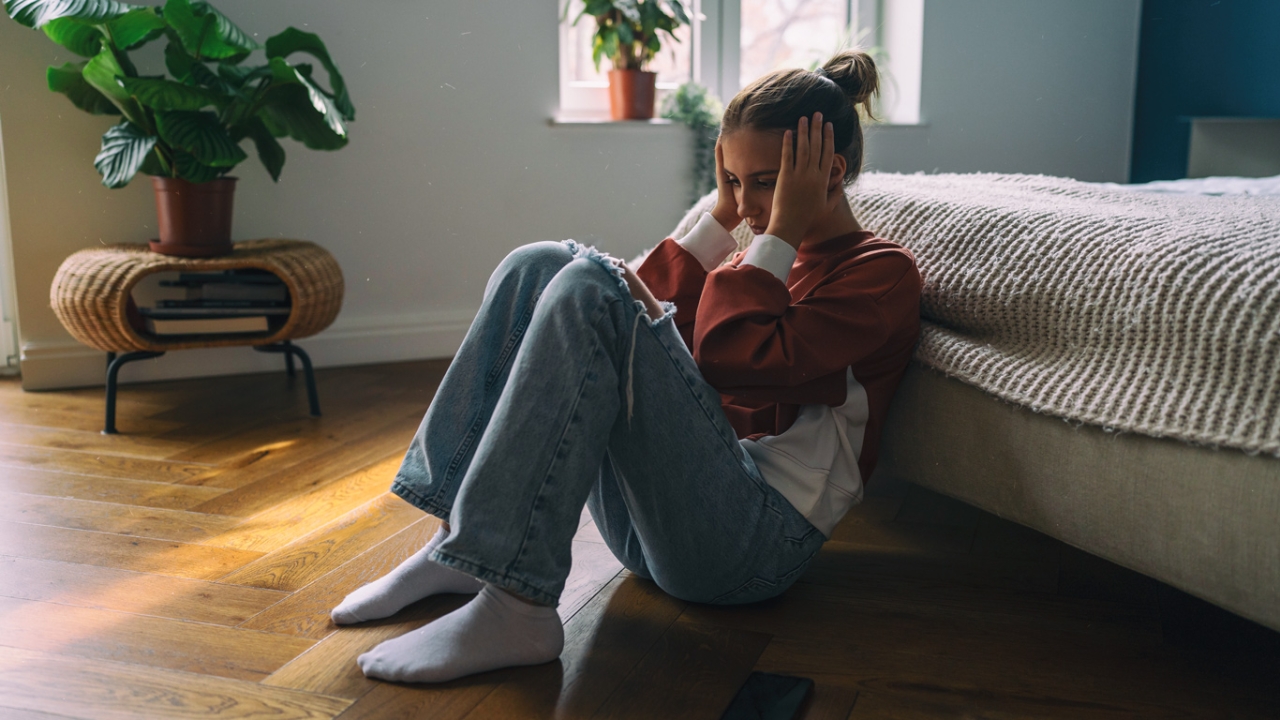 Eine junge Frau sitzt mit angezogenen Beinen auf dem Boden und hat ihre Hände am Kopf