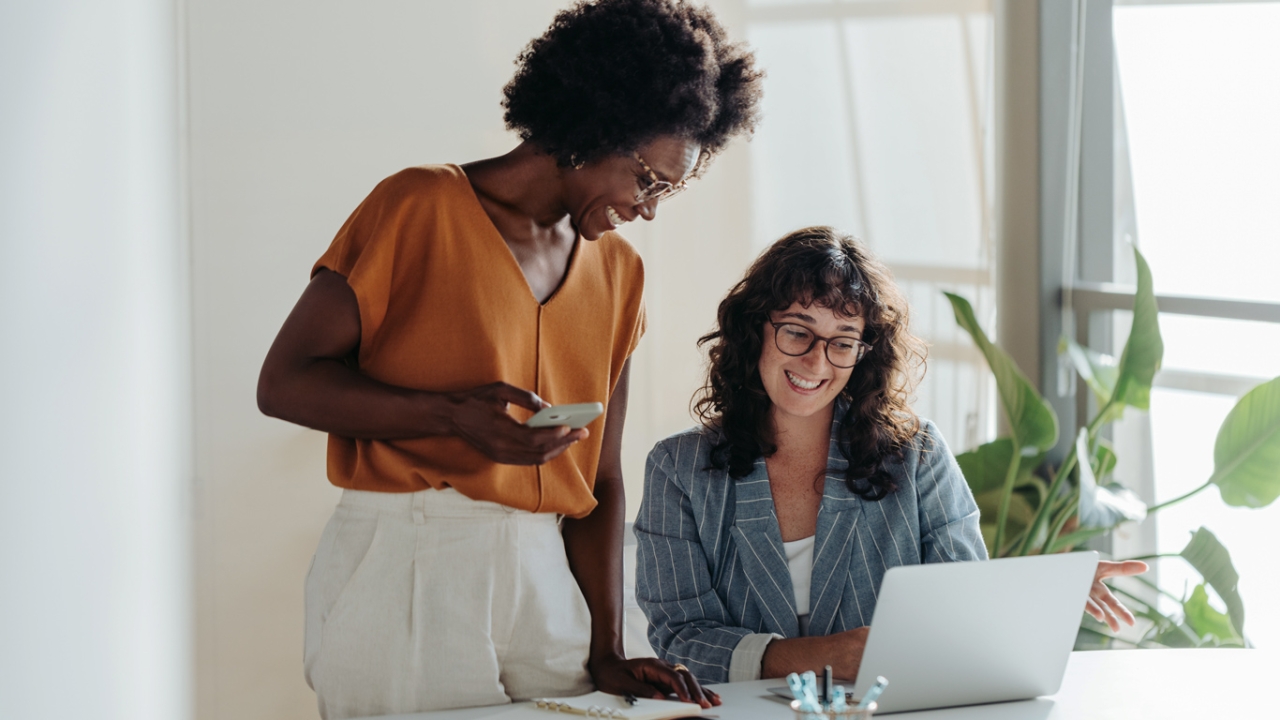Stress auf der Arbeit: Tipps und Übungen für einen entspannten Arbeitsalltag Zwei berufstätige Frauen lächeln und arbeiten gemeinsam an einem Laptop in einem hellen Büro.