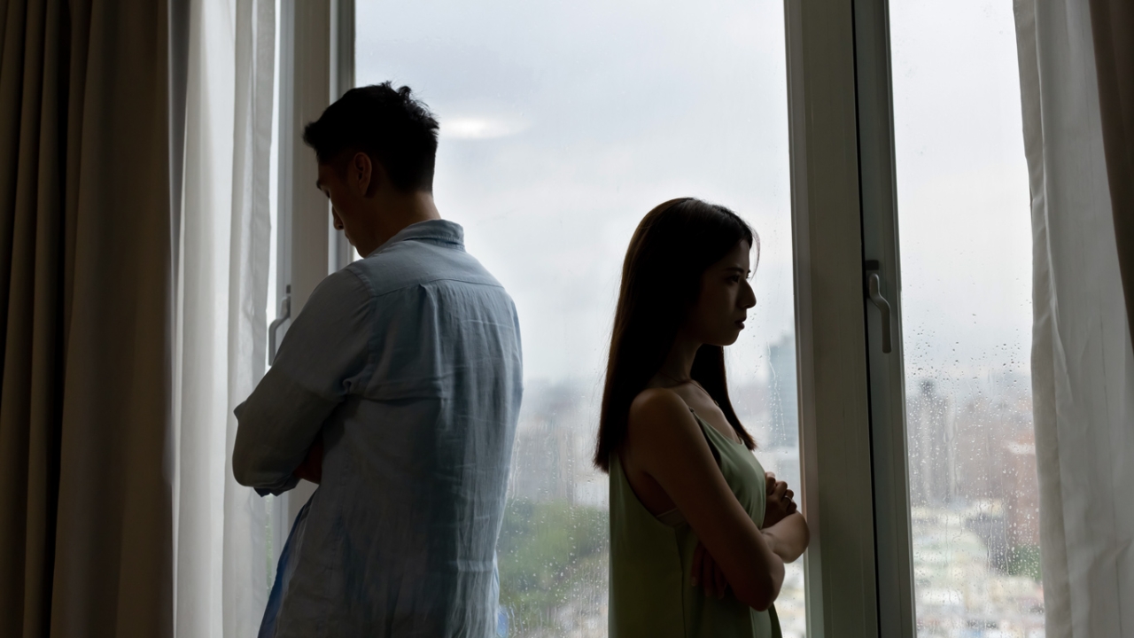 Ein Mann und eine Frau stehen abgewandt voneinander vor einem Fenster, eine sichtbare Spannung zwischen ihnen. Trennung nach langer Beziehung