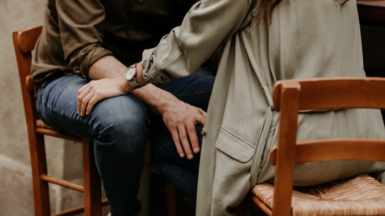 Zwei Personen sitzen nah beieinander auf Holzstühlen, wobei eine Person die Hand fürsorglich auf das Knie der anderen legt. | 12 Beziehungsfragen