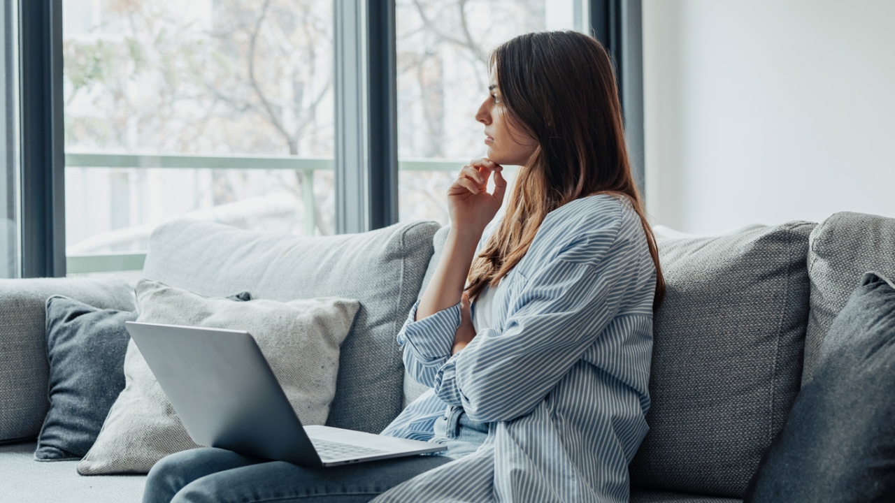 Eine Frau sitzt mit einem Laptop auf einem Sofa und schaut nachdenklich aus dem Fenster. | Schlagfertigkeit lernen