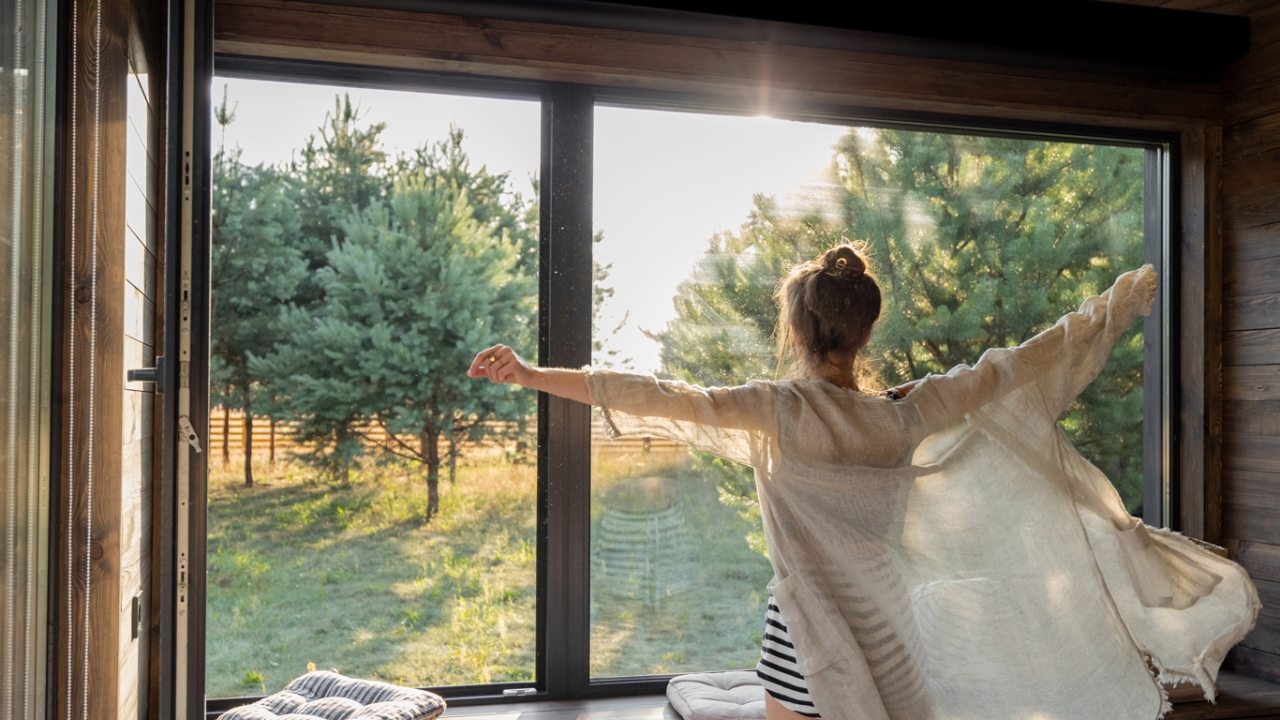 Eine Person mit ausgebreiteten Armen steht vor einem großen Fenster und genießt den Blick auf einen sonnigen Garten mit Bäumen.