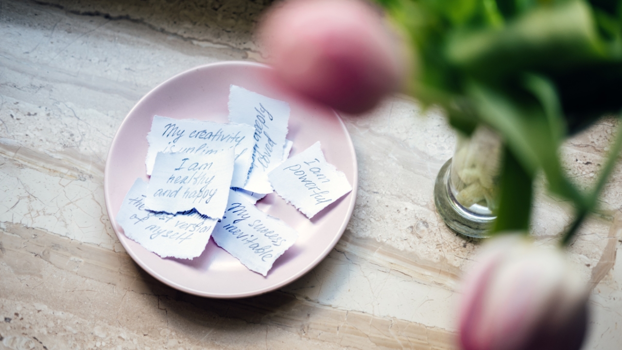 Was sind Affirmationen? Wirkung und Beispiele für jeden Tag Ein rosa Teller mit handgeschriebenen Affirmationen auf zerrissenen Zetteln, daneben eine Vase mit Tulpen.