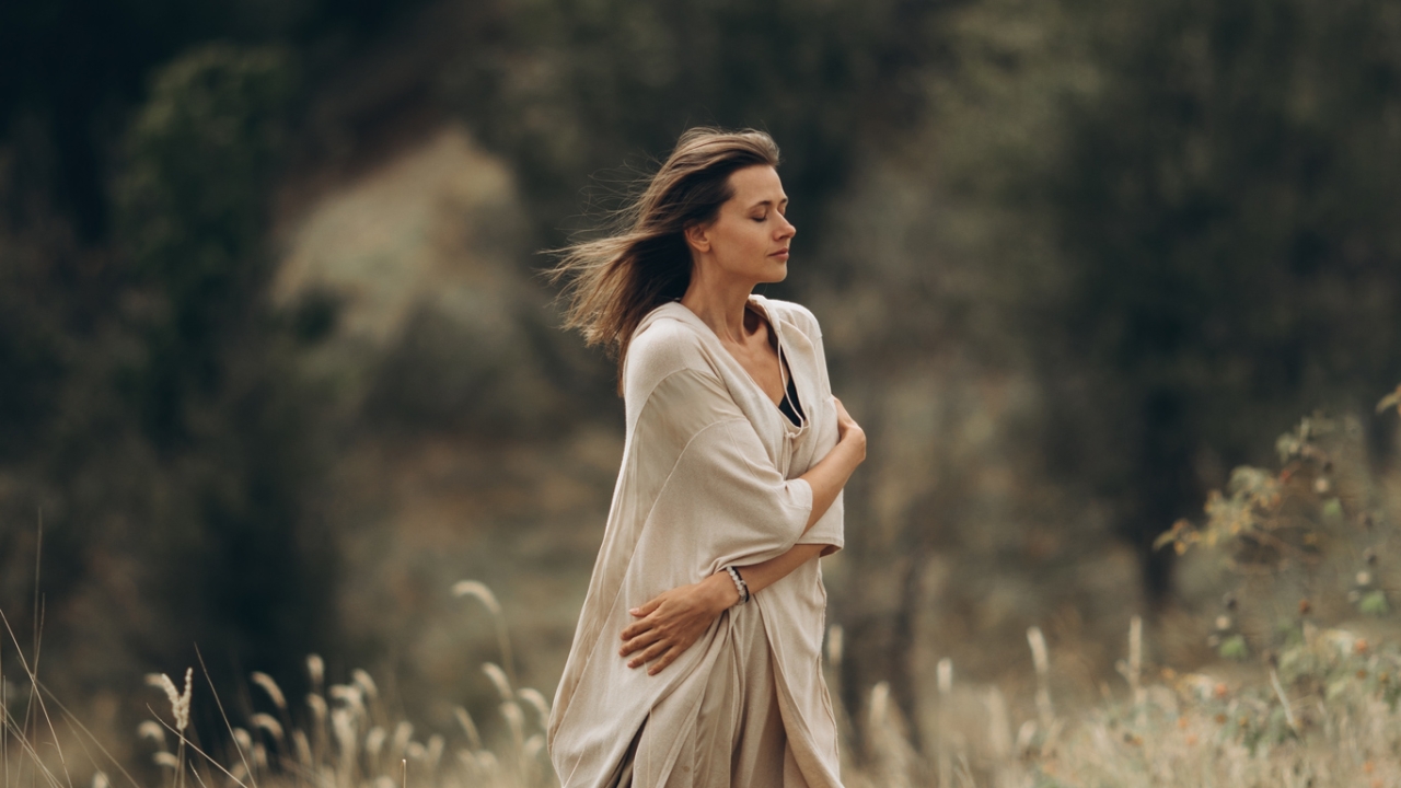 Eine Frau mit geschlossenen Augen steht in der Natur und hält ihre Arme um sich selbst geschlungen, während der Wind ihr Haar weht. | Spiritual Bypassing