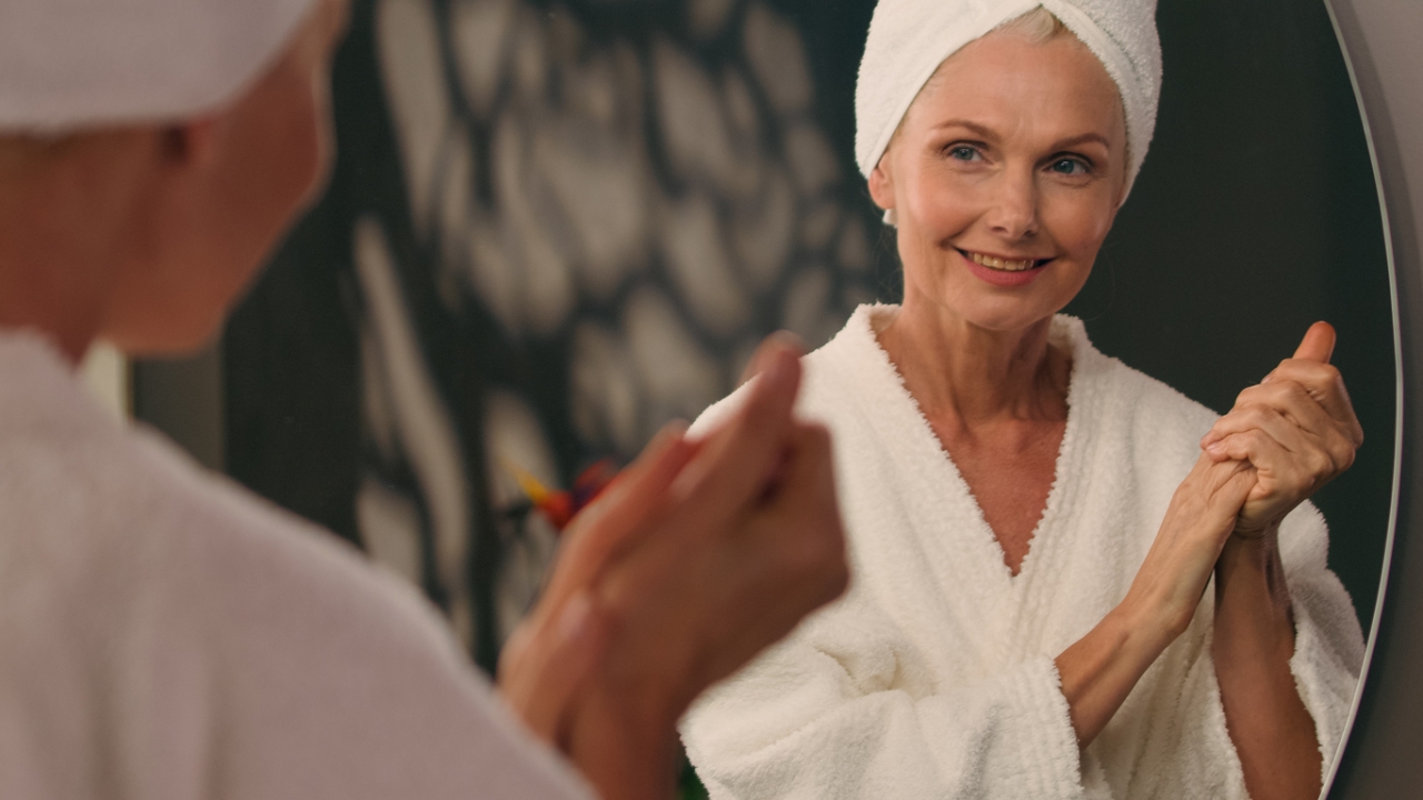 Eine Frau in Bademantel und Handtuchturban blickt lächelnd in den Spiegel | Angst vor dem Älterwerden