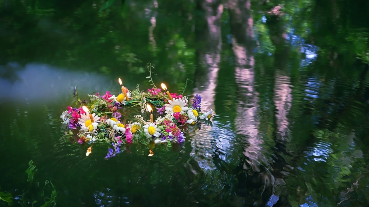 Jahreskreisfeste: Ein bunter Blumenkranz mit brennenden Kerzen schwimmt auf ruhigem Wasser im Wald.
