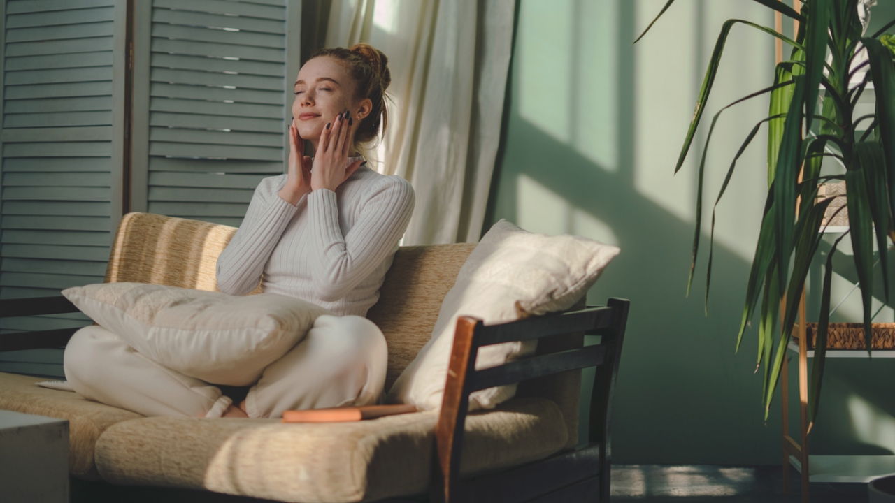 Lymphe anregen: Eine Frau sitzt entspannt im Schneidersitz auf einem Sofa und massiert sanft ihr Gesicht.