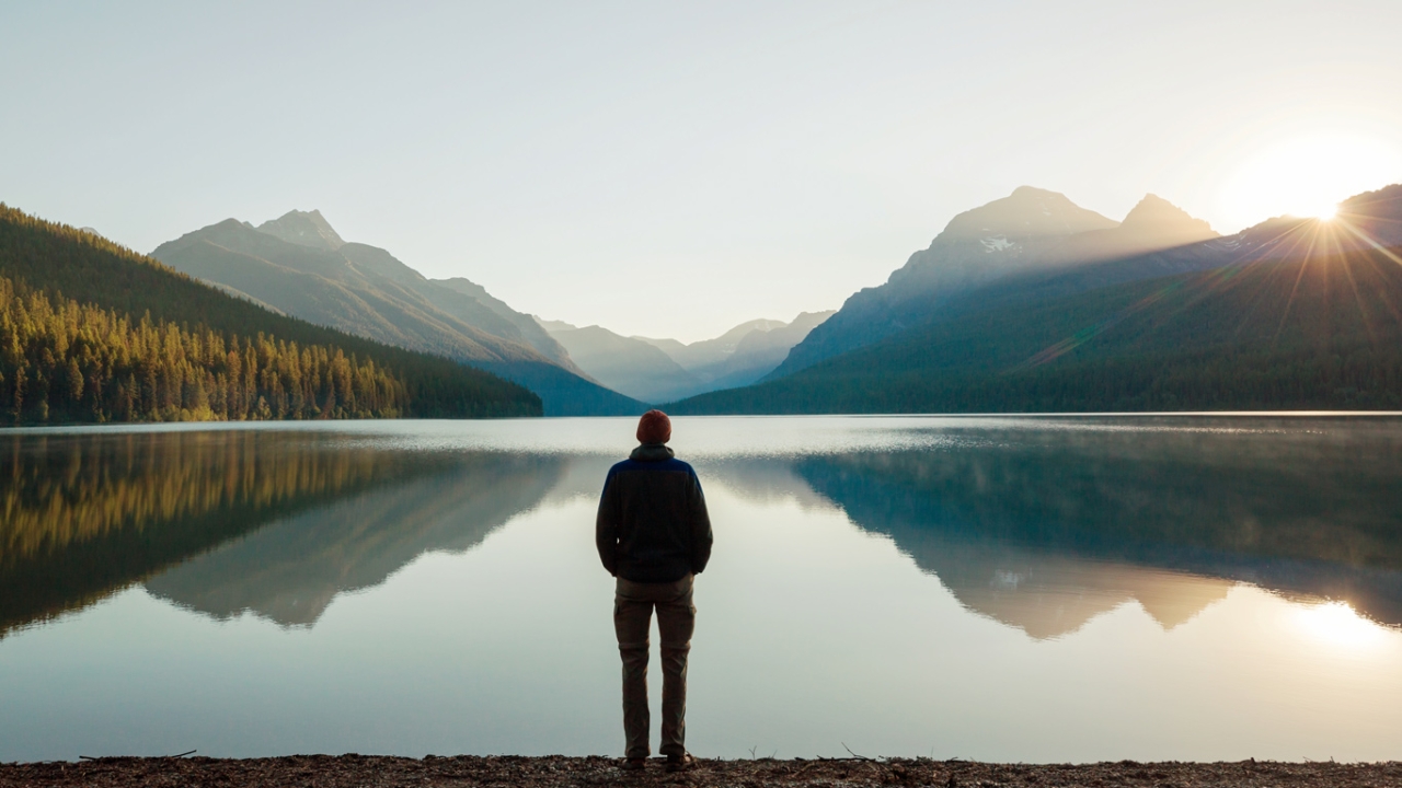 Schwere Zeiten überstehen: Eine Person steht am Ufer eines stillen Bergsees und blickt in den Sonnenaufgang über den Bergen
