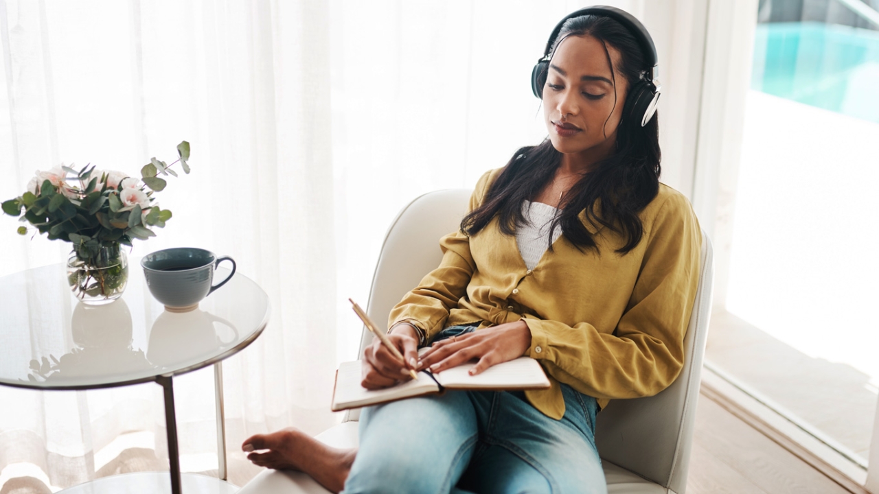 Journaling: Ideen und Fragen für dein persönliches Tagebuch Journaling: Eine Frau sitzt entspannt mit Kopfhörern in einem Sessel und schreibt in ein Journal.