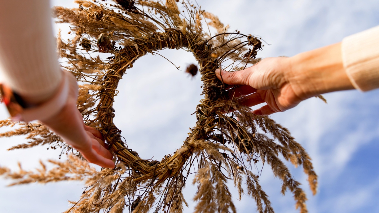 Lughnasadh: Zwei Hände halten einen dekorativen Kranz aus getrocknetem Getreide vor einem hellen Himmel.