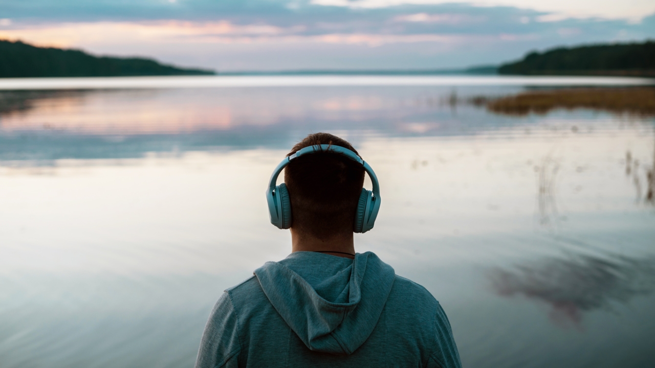 Ein Mann von hinten gesehen trägt Kopfhörer und blickt über einen ruhigen See bei Sonnenuntergang