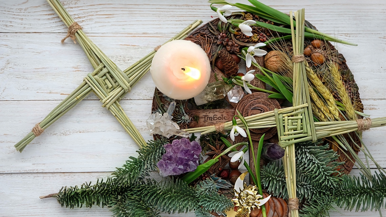 Ein Arrangement zur Feier von Imbolc mit einer brennenden Kerze, umgeben von zwei geflochtenen Brigidskreuzen aus Stroh, Tannenzweigen, Schneeglöckchen und verschiedenen Naturmaterialien.