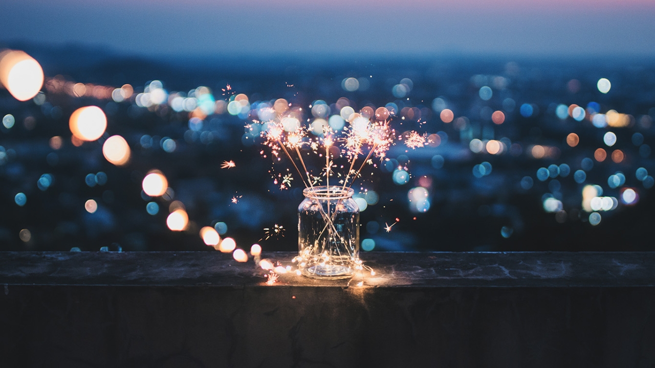 Ein Einmachglas mit brennenden Wunderkerzen steht auf einer Brüstung vor dem unscharfen, nachtblauen Lichtermeer einer Stadt. Die Szene vermittelt eine gemütliche Silvesterstimmung.