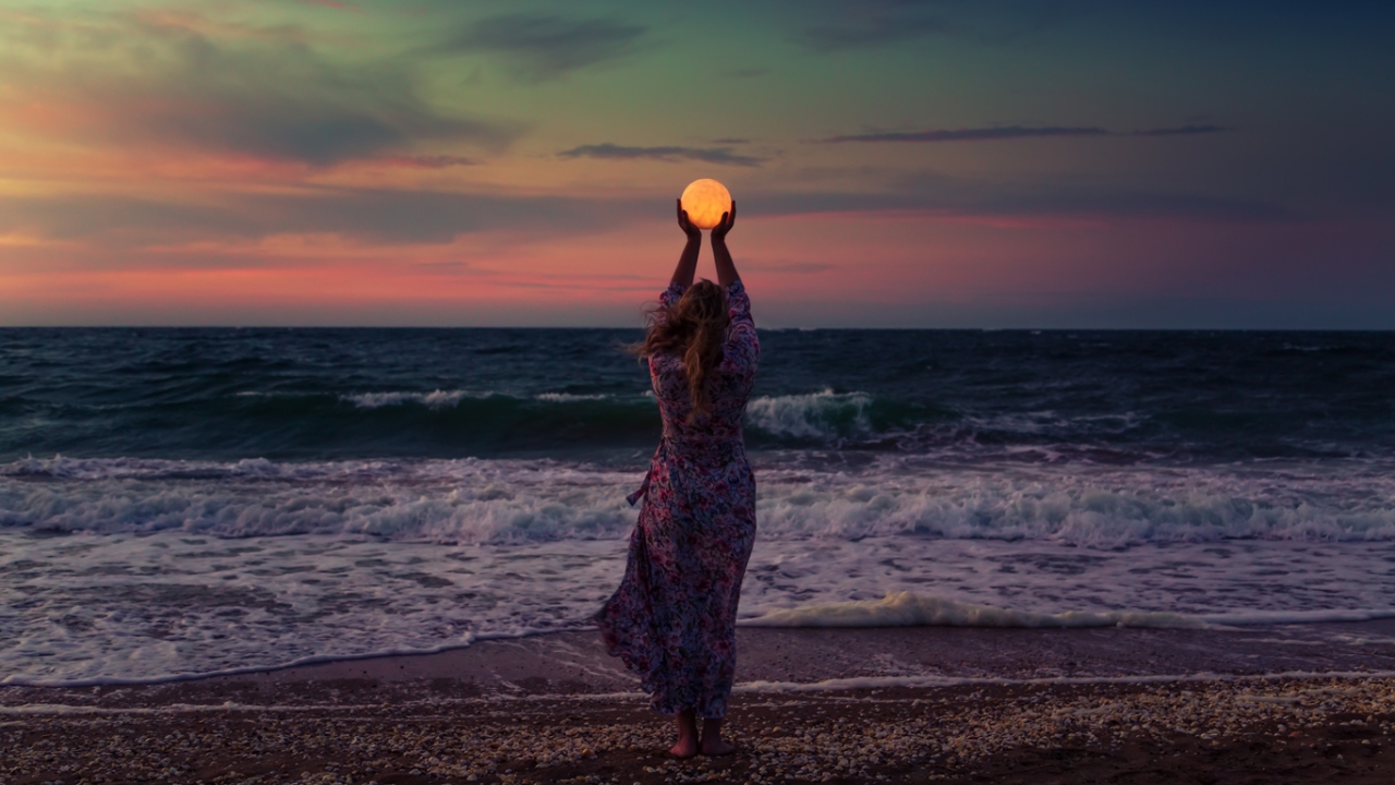 Rückansicht einer Frau im Kleid am Strand, die vor dem Vollmond ihre Arme emporstreckt | Vollmondrituale.