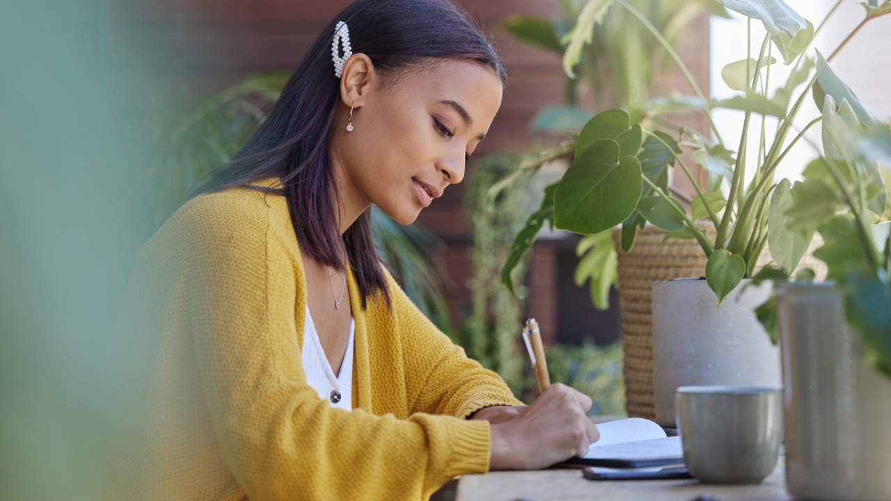 Frau in gelber Strickjacke die an einem Schreibtisch sitzt und in ein Notizheft schreibt. | Bucket List Ideen